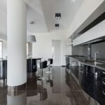Μοντέρνα μαύρη κουζίνα με γυαλιστερά ντουλάπια, υψηλές καρέκλες και ενσωματωμένο φωτισμό οροφής.