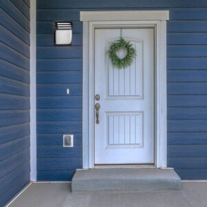 Φωτιστικό εξωτερικού χώρου σε φωτεινό λευκό και μαύρο, τοποθετημένο δίπλα σε λευκή εξώπορτα με γαλάζιο φόντο.